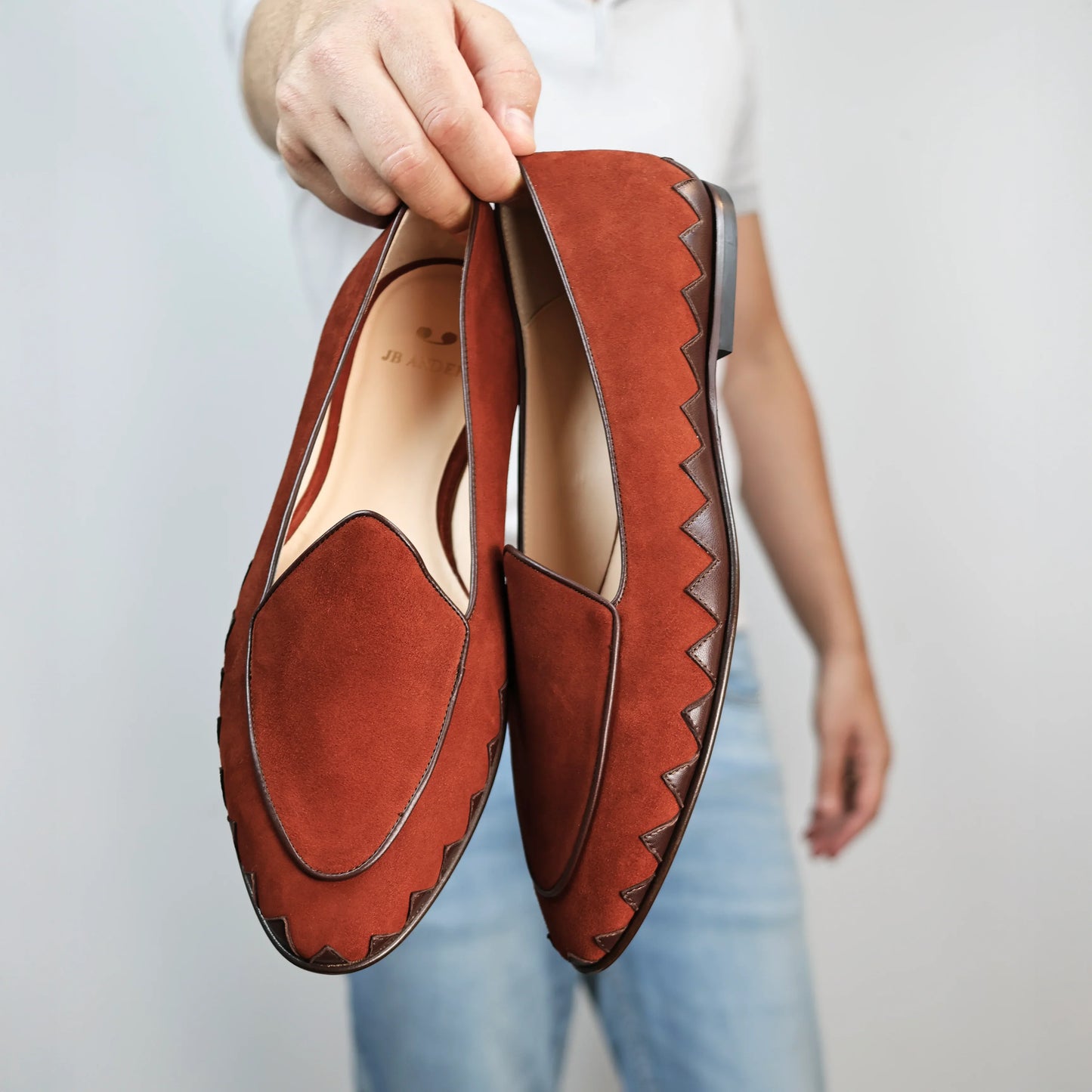 Model holding JB Ander men's Great White smoking slipper loafers in SoHo Sunset burnt umber suede with unique leather trim, and handmade Italian craftsmanship.