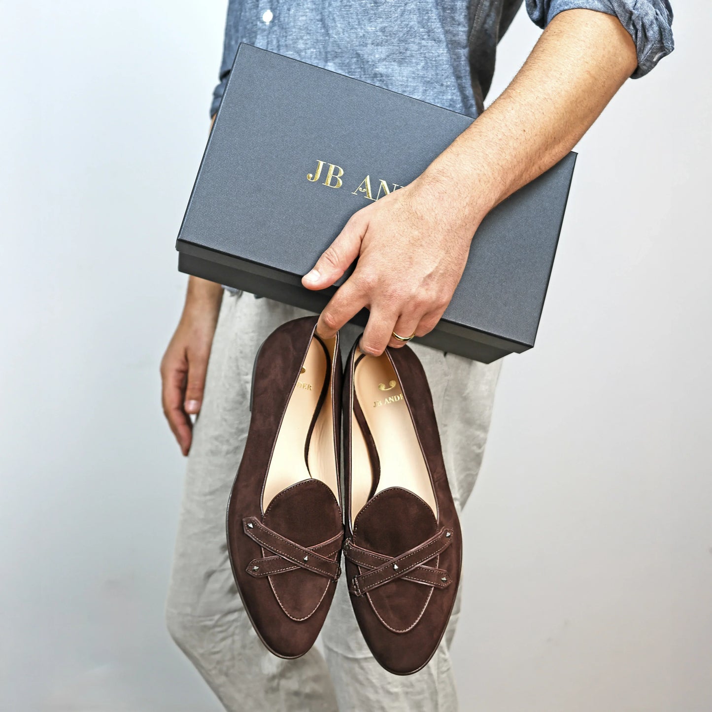 Model holding JB Ander's Lincoln saddle loafer in brown Italian suede – handcrafted in Italy with dual leather straps on vamp, antiqued gold hardware, and luxurious calfskin insole.