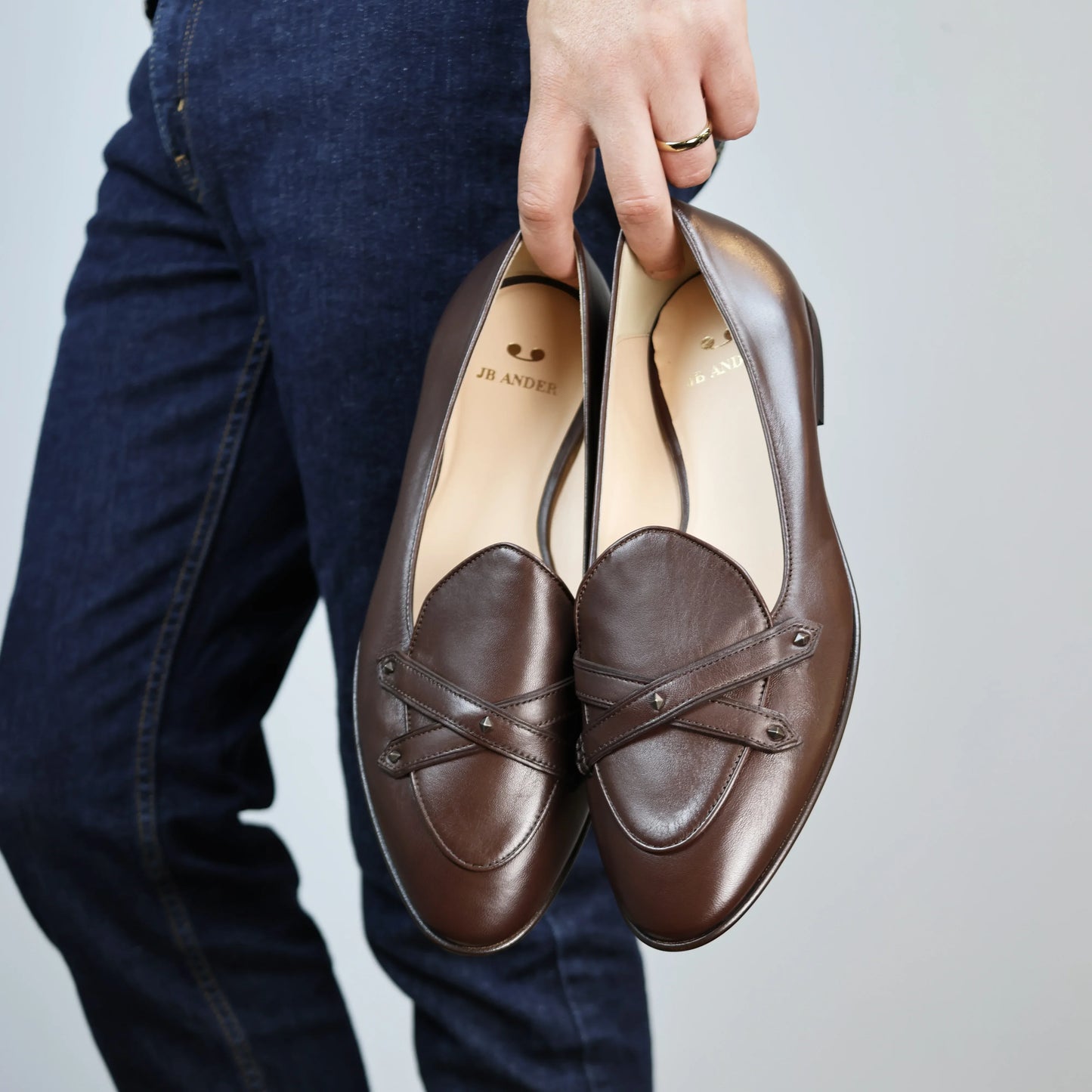 Model holding JB Ander's Lincoln saddle loafer in chestnut brown Italian leather – handcrafted in Italy with dual straps on vamp, antiqued gold hardware, and luxurious calfskin insole.