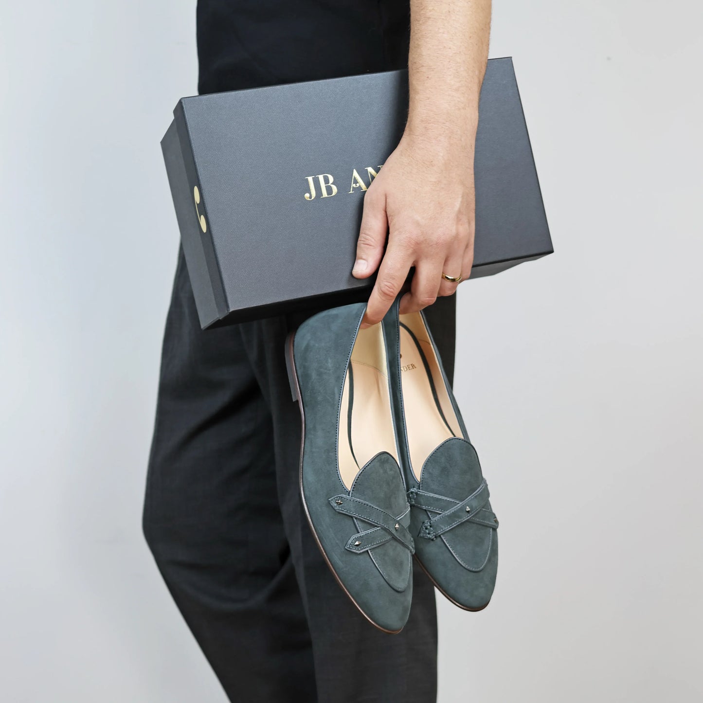 Model holding JB Ander's Lincoln saddle loafer in emerald green Italian suede – handcrafted in Italy with dual leather straps on vamp, antiqued gold hardware, and luxurious calfskin insole.