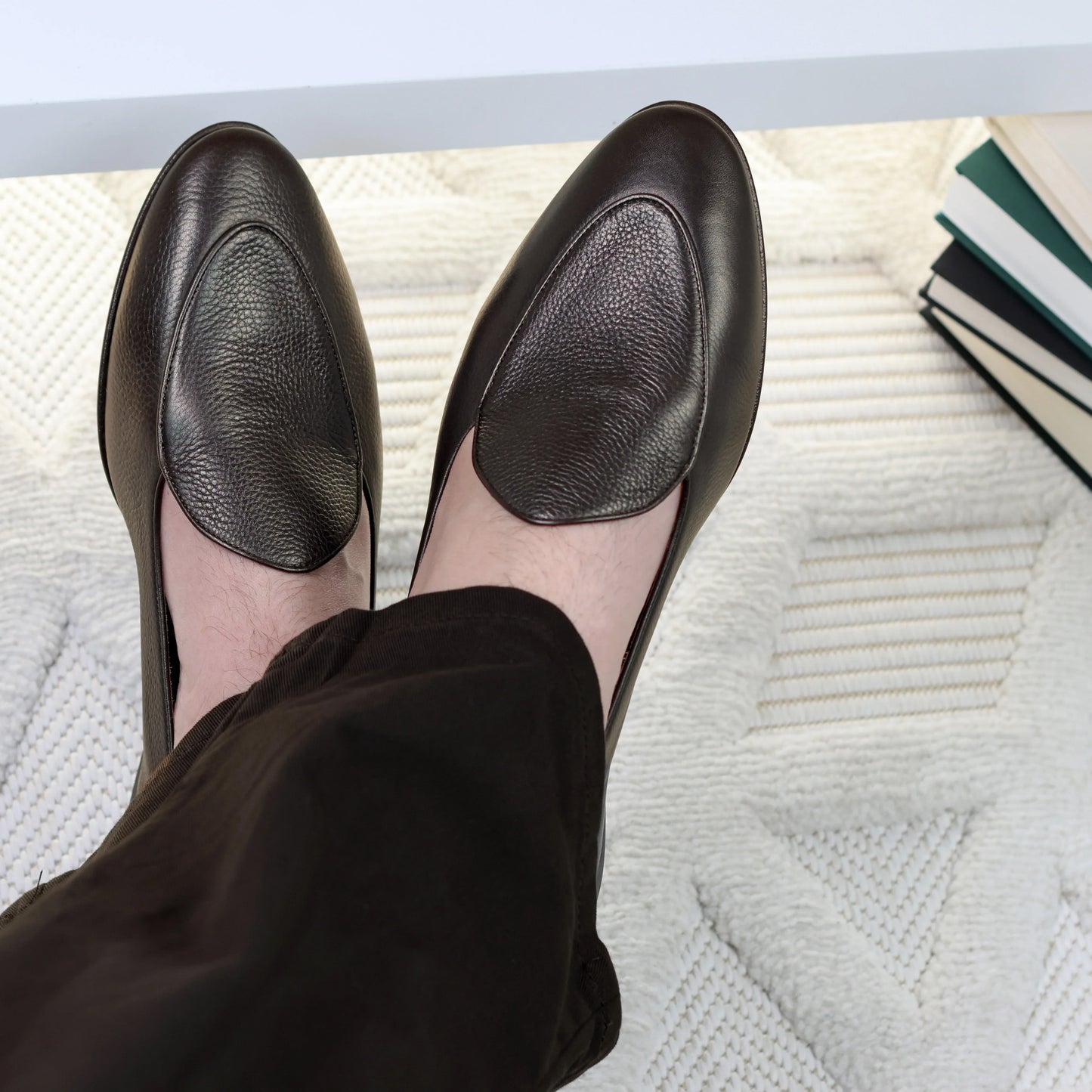 Person wearing JB Ander leather shoes on a textured surface with books in the background