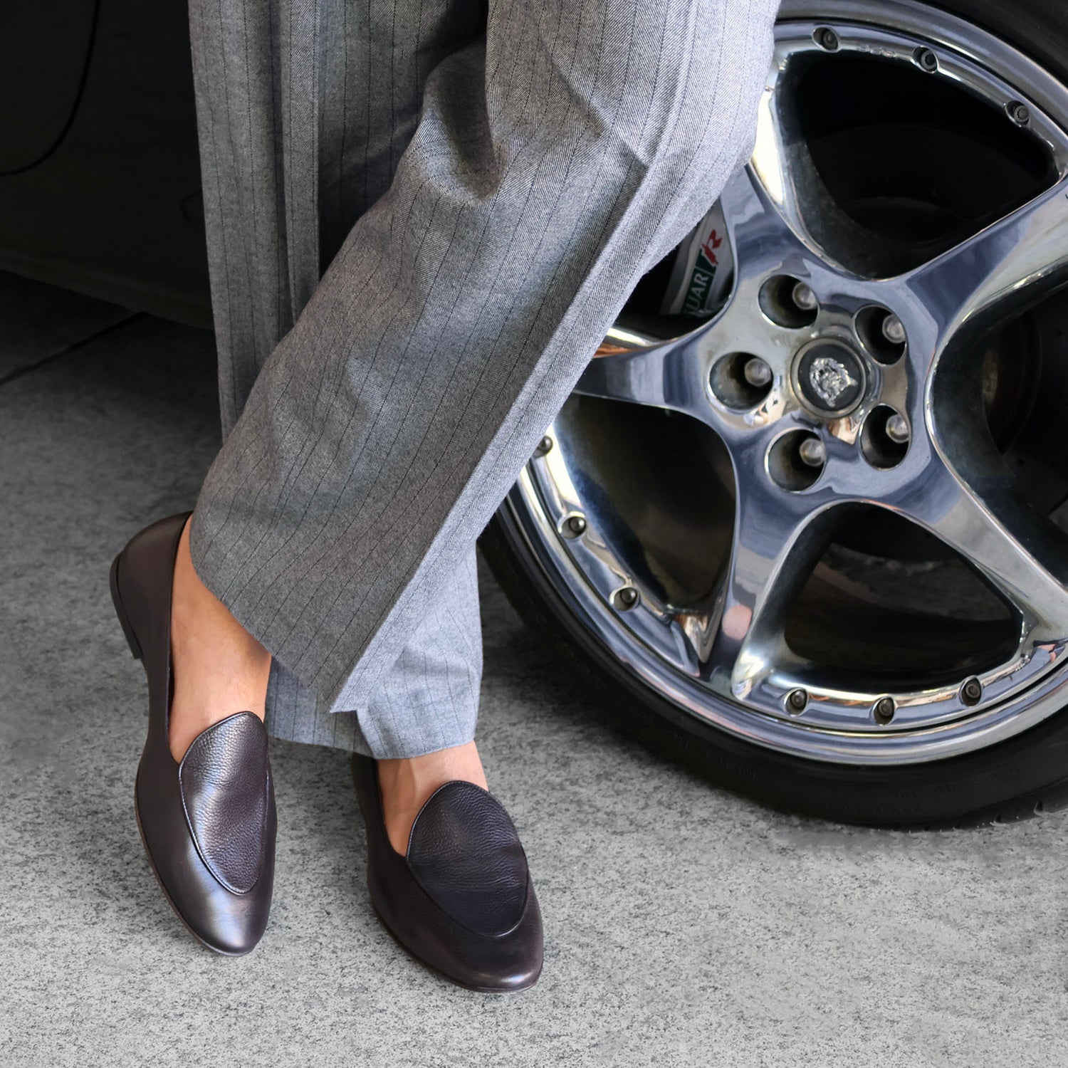 Model wearing gray pants and JB Ander espresso brown pebbled leather Venetian loafers standing next to a car wheel.