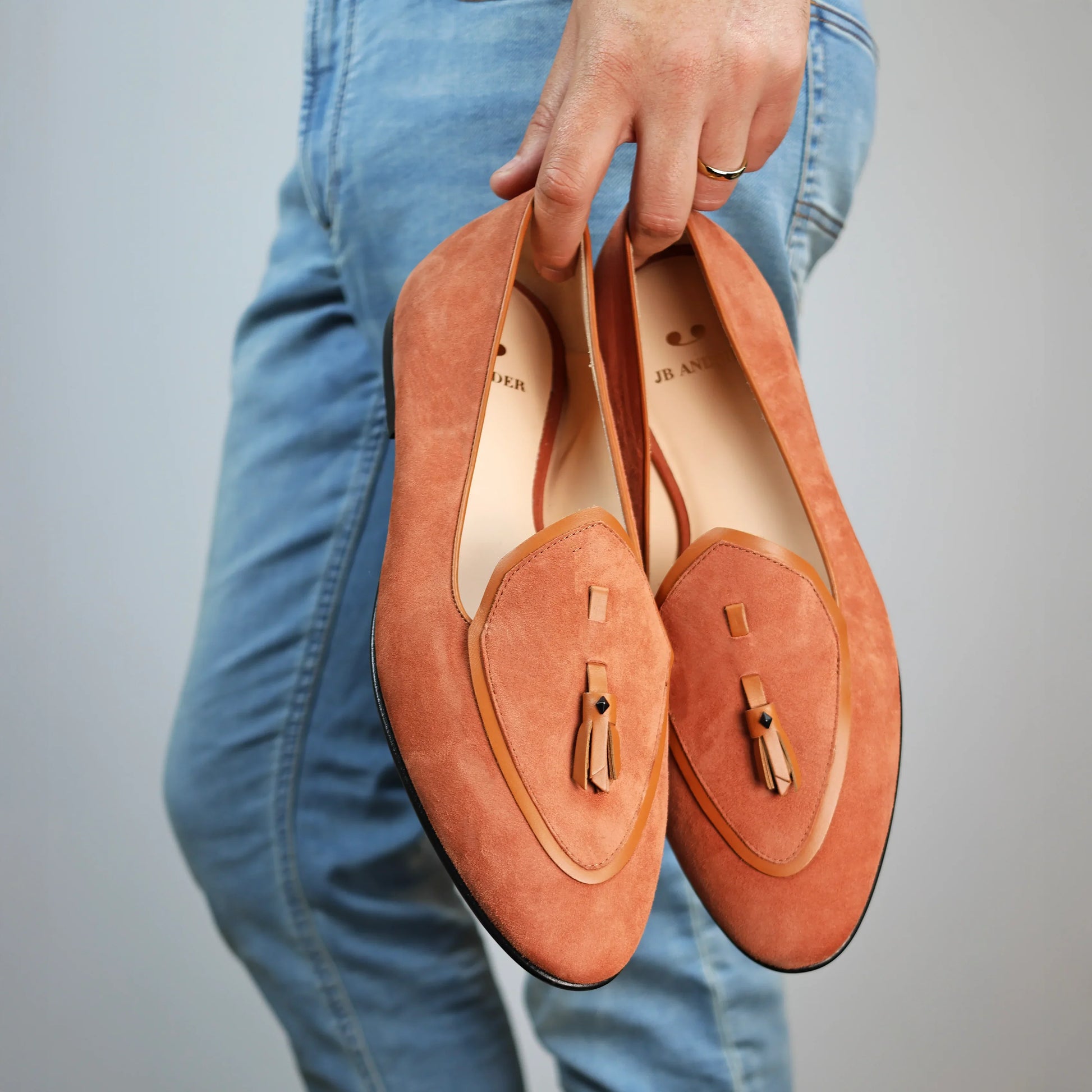 Model holding JB Ander men's Valedictorian tassel loafers in terracotta suede with supple leather trim, and handmade Italian craftsmanship.