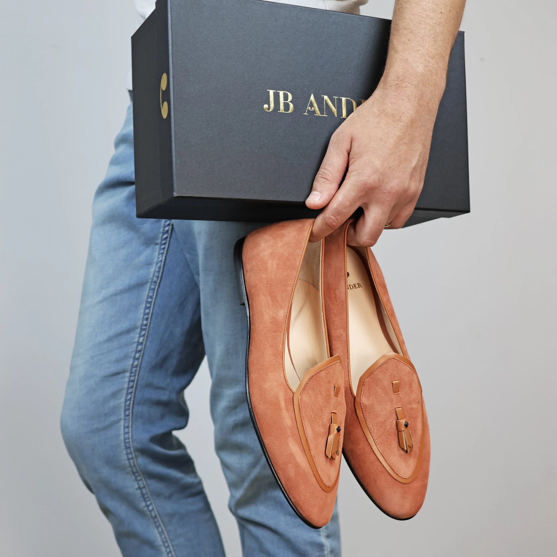 Model holding JB Ander men's Valedictorian tassel loafers in terracotta suede with supple leather trim, and handmade Italian craftsmanship.