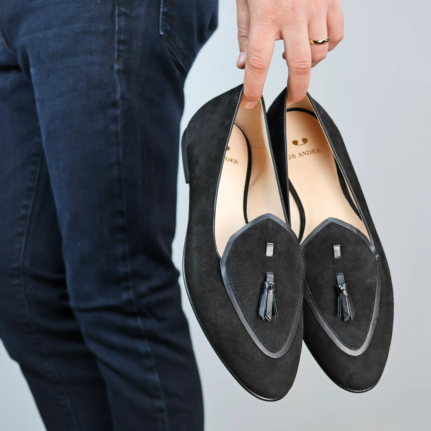 Model holding JB Ander men's Valedictorian tassel loafers in Nero black suede with black leather trim, and handmade Italian craftsmanship.