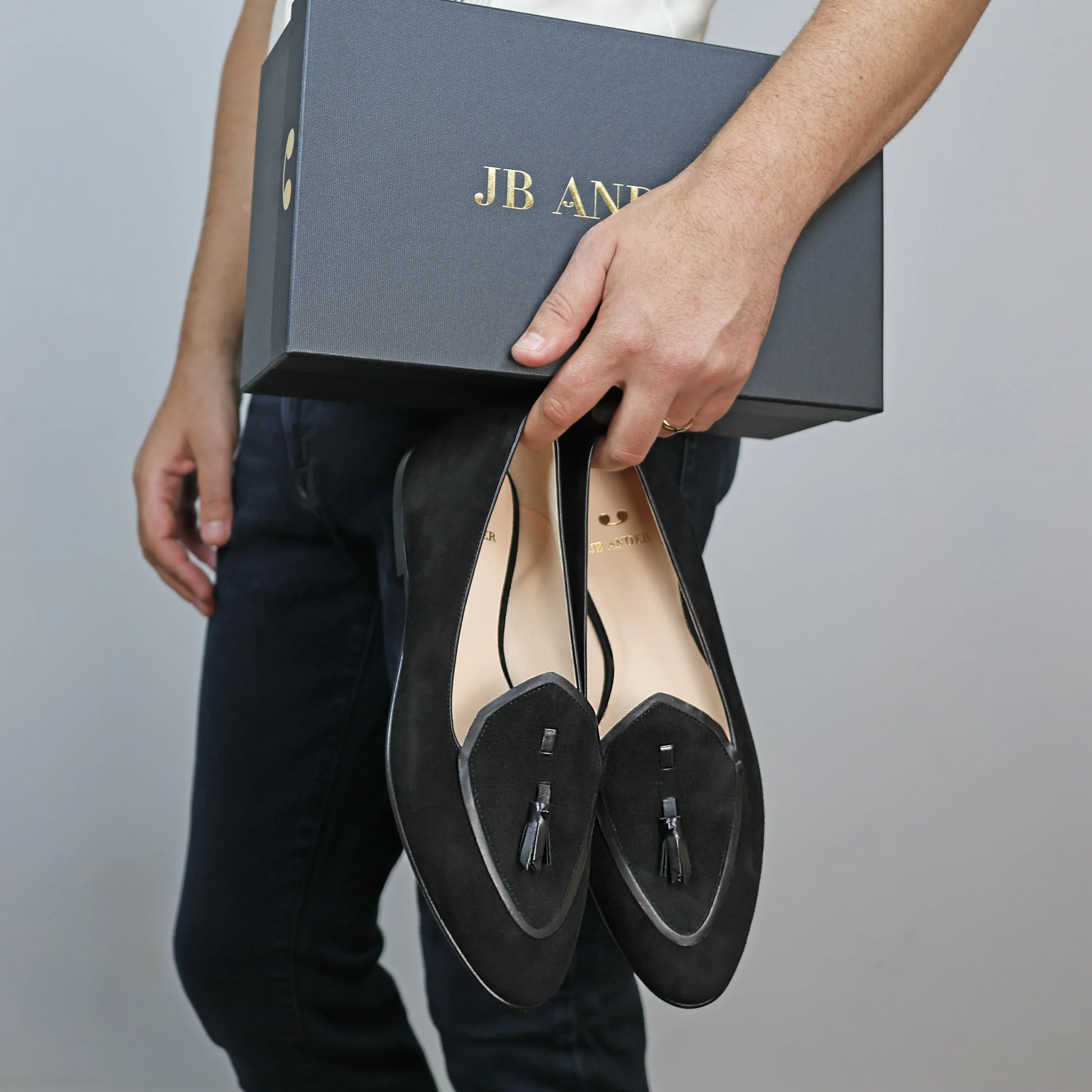 Model holding JB Ander men's Valedictorian tassel loafers in Nero black suede with black leather trim, and handmade Italian craftsmanship.