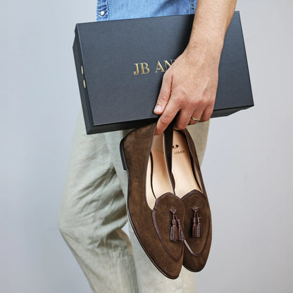 Model holding JB Ander men's Valedictorian tassel loafers in chestnut brown suede with supple leather trim, and handmade Italian craftsmanship.