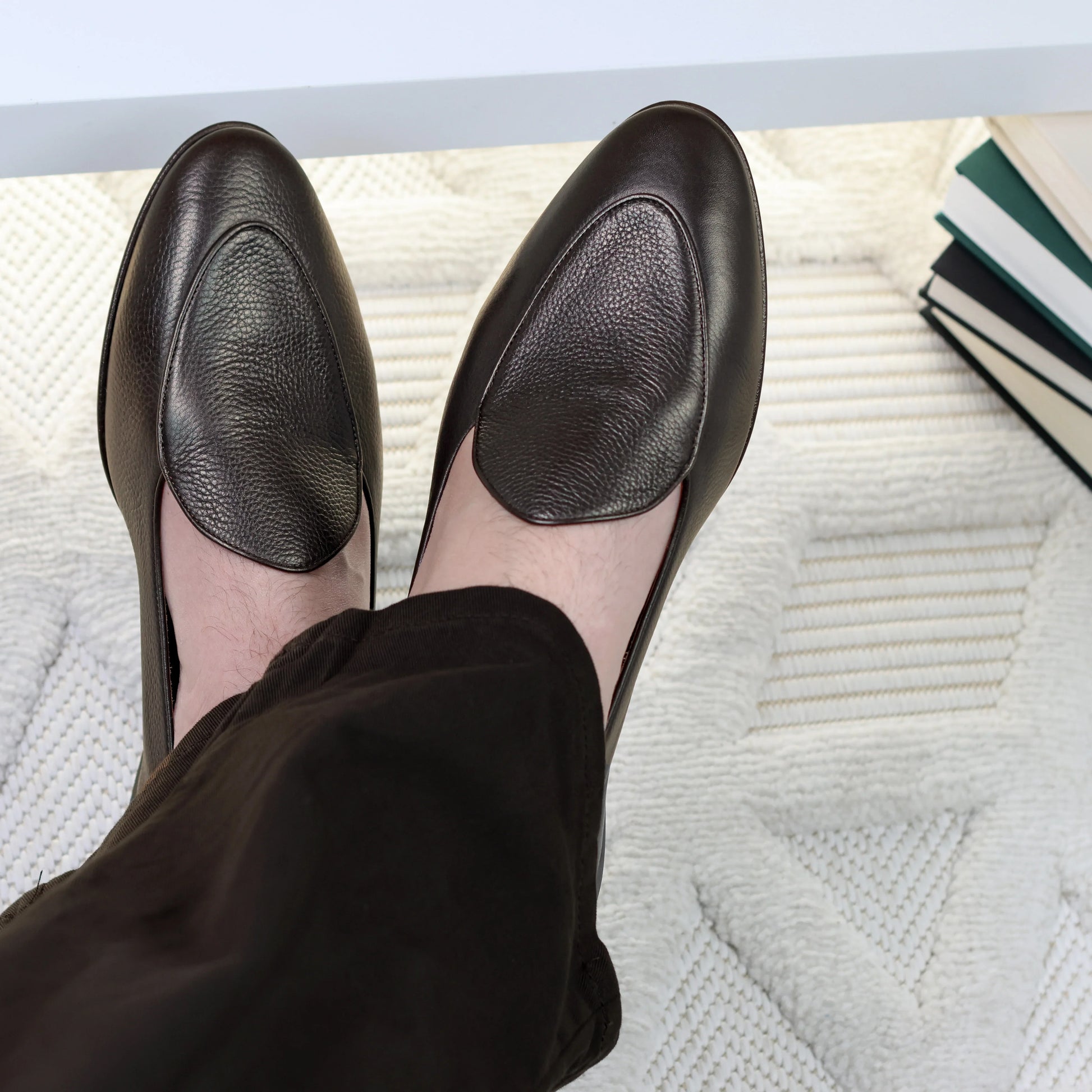 Person wearing JB Ander leather shoes on a textured surface with books in the background